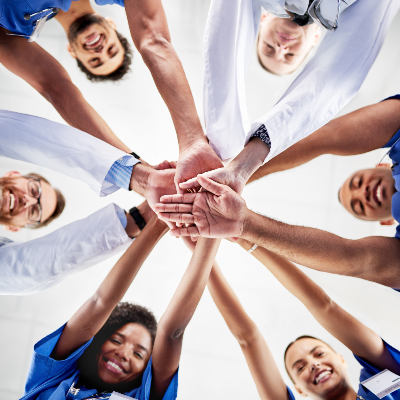 Group of smiling medical professionals stood in a circle facing inwards towards each other, overlapping hands in the centre of the circle.