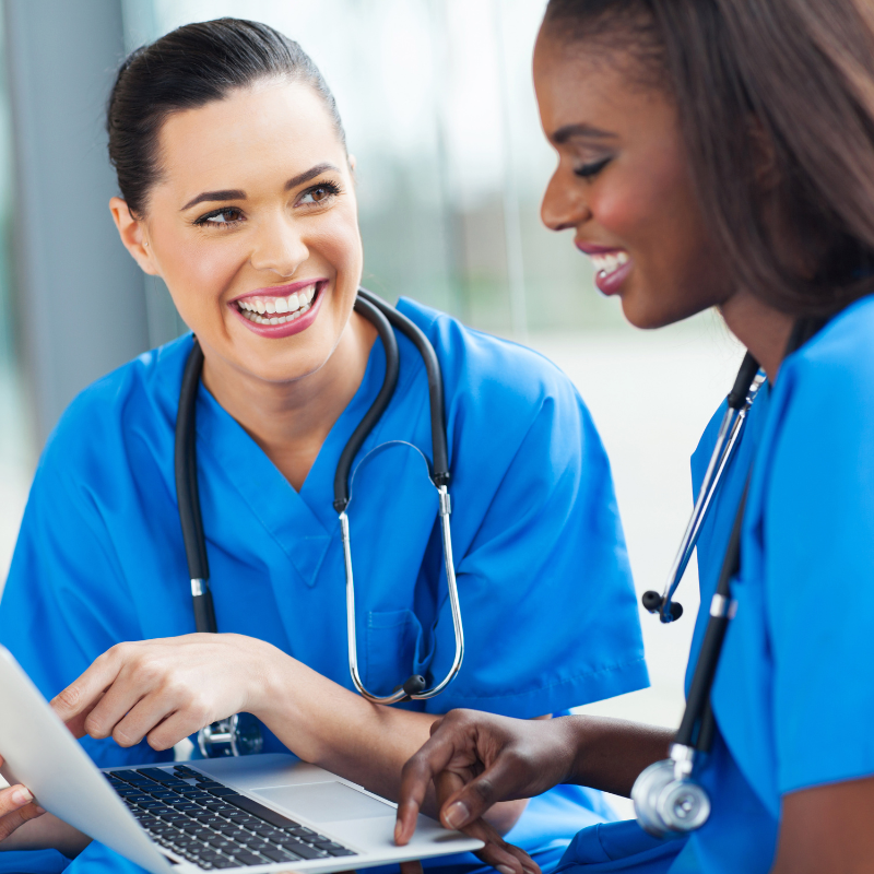 Two smiling female medical professionals with stethoscopes around their necks, looking at a laptop.